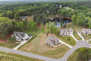 Aerial view of a residential area featuring several houses with driveways, surrounded by greenery and trees. A small lake is visible in the background, and a road curves through the neighborhood.