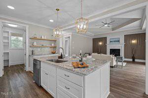 Modern kitchen and living area with wooden floors. The kitchen features white cabinets, a granite countertop with a sink, and pendant lights. Open shelves hold dishes. The living area has a brown accent wall and fireplace, with a ceiling fan above.