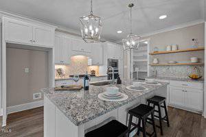 Modern kitchen with white cabinets and a granite island featuring three bar stools. Pendant lights hang above the island, which is set with wine glasses and plates. Open shelves display dishes, and a tiled backsplash is visible behind the stove.