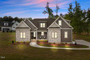 A two-story house with a modern design is illuminated at dusk. The exterior features light brick and dark siding accents. A curved driveway leads to the entrance. The sky in the background is colorful with shades of pink and blue.