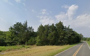 A rural road runs alongside a dense line of trees under a partly cloudy sky. The two-lane asphalt road has no markings, and the grass shoulder is trimmed. The surrounding area appears quiet and natural.