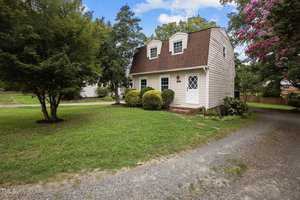 Nestled among trees and shrubs, this charming two-story house on 209 Park Avenue in Creedmoor features a gambrel roof and white siding. A gravel driveway leads to a grassy yard, where a small path welcomes you to the elegant white door.