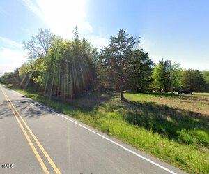 A rural road bordered by trees and a grassy area on a sunny day. Sunlight shines through the tree branches, creating bright rays across the scene. The road has a double yellow line, and there are scattered bushes and open space beside it.