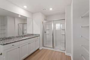 A modern bathroom featuring a granite-topped vanity with a single sink and a large mirror above. Next to it is a glass-enclosed shower with white tile. The room has white walls, recessed lighting, and wooden flooring. Wire shelves are visible on the right.