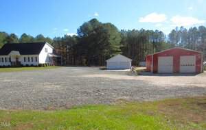 A white two-story house with a porch is on the left, surrounded by a large gravel area. To the right, there is a red barn-like structure with two garage doors. A smaller white garage is between them. In the background, there is a dense forest.