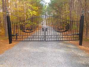 A closed wrought iron gate with artistic deer and tree patterns stands at the entrance of a gravel driveway. The gate is flanked by tall trees on both sides.