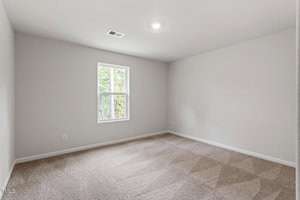 An empty room with beige carpet, white walls, and a single window. A ceiling light is turned on, illuminating the space. The window offers a view of some trees outside.