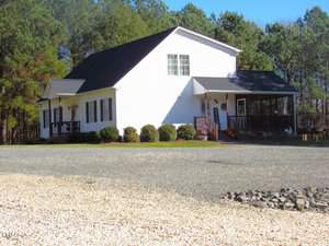 A white house with a dark roof is surrounded by trees. The house features a front porch, windows with shutters, and a gravel driveway extending in front. It's a sunny day with a clear blue sky.