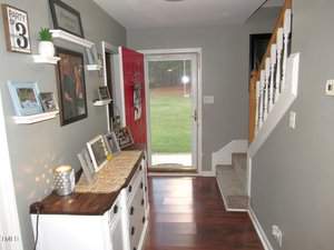 A hallway with a wooden floor, white dresser, and a red front door opening to a grassy yard. The walls display framed photos and a sign. A staircase with a white railing leads upstairs. A lit candle sits on the dresser next to small decor items.