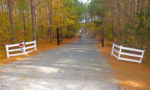 A gravel driveway leads to a closed black metal gate surrounded by a forest of tall pine trees. On both sides of the driveway, there are white wooden fences and a security keypad on the left. The scene is lit by daylight.