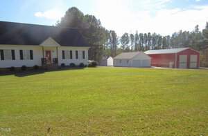 A landscape view shows a white single-story house with a dark roof on the left, a large neatly mowed lawn in front, and a red barn with white doors on the right. A small white shed is located between the house and barn. Trees are in the background.