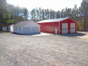 A red metal barn with two large garage doors is situated next to a smaller white building with two garage doors. Both structures are surrounded by a gravel area and trees in the background.