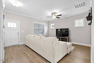 A modern living room with a large beige sectional sofa facing a flat-screen TV on a wooden stand. The room features wood flooring, a ceiling fan, and two windows with white blinds. The walls are painted light gray, and there's a white entrance door.