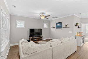 A modern living room with light gray walls, a beige sectional sofa, and a flat-screen TV on a wooden stand. The room features wood flooring, a ceiling fan with a light, and a few decorative shelves and framed pictures on the walls.