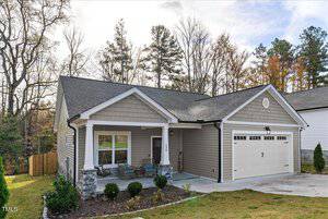 A single-story house with a gray facade, white trim, and a dark roof. The front porch features stone accents and two chairs. The attached garage has a double door. The yard is partially landscaped with shrubs, and there are trees in the background.