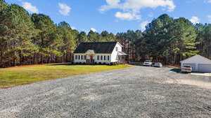 Situated at 3214 Tump Wilkins Road, this white house with a gabled roof sits on a spacious lot surrounded by trees. A large gravel driveway leads to the house, where two vehicles are parked alongside a detached garage. The sky is partly cloudy, adding to the serene atmosphere of this home.