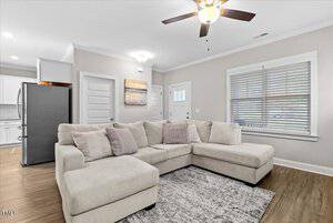 A living room with a light beige sectional sofa, multiple cushions, and a patterned rug. The room features a ceiling fan, a large window with blinds, and artwork on the wall. In the background, a kitchen with white cabinets and a stainless steel fridge is visible.