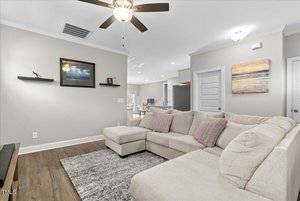 A cozy living room with a large beige sectional sofa and a gray rug on wooden flooring. The walls are light gray, adorned with two paintings and a small shelf. A ceiling fan with a light is above, and a partially visible kitchen is in the background.