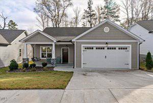 A modern single-story house at 134 Kipling Drive, complete with a gabled roof and inviting front porch. The facade features gray siding accented by white trim, while a two-car garage with white doors complements the design. Nestled in Oxford, it boasts a landscaped area with shrubs and concrete driveway.