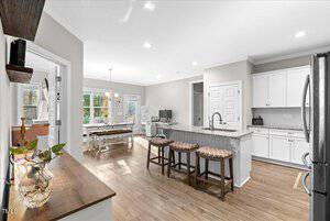 A spacious kitchen with light wood flooring and white cabinets. A granite island with three bar stools is central. To the left is a dining area with a table and chairs. Natural light streams through large windows, illuminating the space.