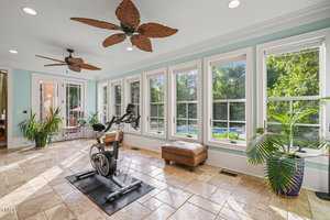 A bright room with large windows showing a greenery view. There is an exercise bike on a mat facing the windows. The room features tile flooring, ceiling fans with leaf-shaped blades, a brown ottoman, and potted plants.