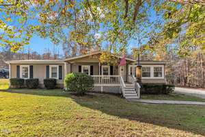 Nestled in the serene surroundings of 1619 Hunters Trail, Creedmoor, this charming single-story house features beige siding and a welcoming front porch amid lush trees. A pink flag adorns the entrance. The yard is grassy and well-maintained, with shrubs flanking the steps under a clear blue sky.