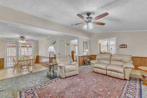 A cozy living room features a beige recliner and sofa on a patterned rug. A ceiling fan is overhead. The space opens to a dining area with a wooden table and chairs, both adorned with ceiling fans. Walls are light, and there's wood paneling halfway up.