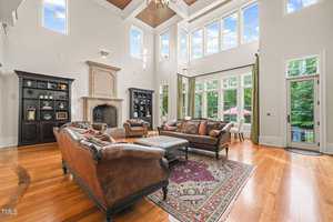 Spacious living room at 1076 Cabin Creek Road in Oxford, featuring high ceilings and large windows. A decorative fireplace complements the leather sofas and patterned rug. Shelves and a small dining area are visible, with hardwood floors and an outside view of greenery completing the setting.