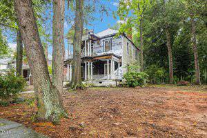 A dilapidated two-story house with peeling paint and broken windows. Surrounded by tall trees and overgrown vegetation, the property shows signs of neglect. The roof appears partially damaged, and the area is littered with debris.