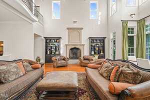 Located at 1076 Cabin Creek Road in Oxford, this spacious living room boasts high ceilings and large windows. It features two brown sofas with patterned cushions, a large ottoman, and two armchairs. A stone fireplace is flanked by dark bookshelves, with hardwood floors and a patterned rug completing the scene.