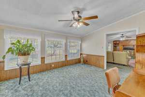 A room with a blue carpet, wooden wainscoting, and large windows. There's a ceiling fan with lights, a potted plant on a small table, and a wooden desk with a chair. An opening leads to a living area with a couch and a fireplace.