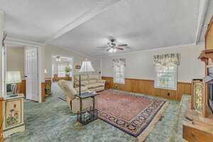 A living room with a blue-green carpet and a large red patterned rug. It features a beige sofa, a floor lamp, and ceiling fans. There is a decorative fireplace on the right, and two windows with floral curtains along one wall. An open doorway leads to another room.