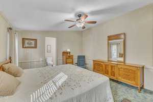 A bedroom featuring a large bed with a patterned quilt, a wooden dresser with a mirror, and a dark reclining chair. An overhead ceiling fan provides light, and an open door reveals a glimpse of a bathroom with a toilet.