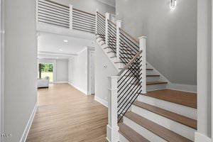 A modern interior features a wooden staircase with metal railings, leading to an upper floor. The space has light-colored walls and wood flooring, with a hallway leading to a room visible through an open doorway.