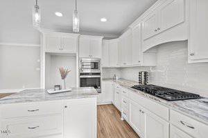 Bright, modern kitchen featuring white cabinetry and marble countertops. The space includes a center island with a vase of dried flowers, a gas cooktop, an integrated oven, and pendant lighting. Light wood flooring completes the design.