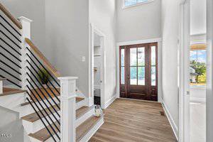 A bright interior entryway featuring a wooden staircase with black metal railings on the left. The room has light gray walls and wood flooring. A dark wooden front door with glass panels lets in natural light. A small potted plant is visible under the stairs.