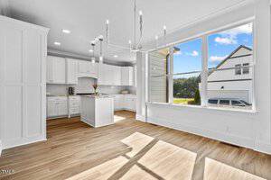 A bright kitchen and dining area with white cabinets, a central island, and a modern chandelier. The space features wood flooring and large windows offering a view of a house and greenery outside.