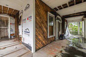 A partially renovated porch with exposed wood and insulation on the walls. The floor shows signs of water damage, and there are large windows. A columned railing is visible in the background, and a tarp covers part of the floor.