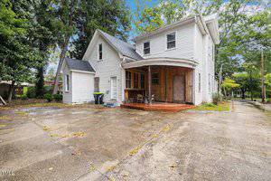 A two-story white house with a covered porch and wooden shutters stands on a concrete driveway. The surrounding area features trees and a quiet street. The sky is partly cloudy.