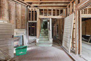 An interior view of an unfinished building showing exposed wooden beams and brick walls. A staircase with a damaged green carpet leads to an upper level, while debris and old doors are scattered on the floor. A green plastic bin is visible on the left.