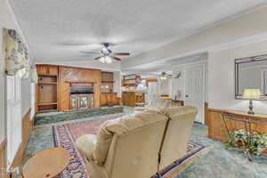 Spacious living room with beige carpet and brown wooden paneling. Features a large beige sofa, wooden coffee table, and a fireplace. Ceiling fans with lights are installed, and a small dining area is visible in the background. A large area rug is centered.