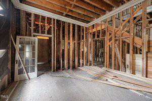 An empty room under renovation with exposed wooden studs and beams, a partially open door, scattered planks on the floor, and visible structural elements.