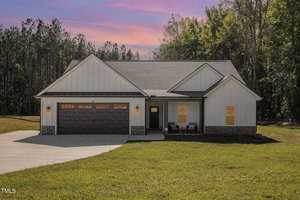 A single-story house on 3656 Bruce Garner Road features white siding and a wooden garage door, beautifully captured at sunset. Located in Wake Forest, the home boasts a small front porch with chairs, surrounded by trees and a grassy lawn.