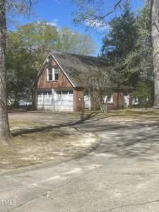 A small, two-story brick garage with white double doors and a pitched roof is surrounded by trees. The driveway curves in front of the building. The sky is clear and blue, and the area is shaded by tree branches.