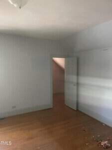 An empty room with light blue walls and a wooden floor. The room features a partially open white door leading to a small space or closet, and there is some debris on the floor near the bottom right corner. A light fixture is visible on the ceiling.