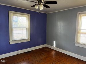 A small, empty room with two windows featuring white blinds. The walls are blue and grey, with a wooden floor. A ceiling fan with lights is installed above. Sunlight filters in through the windows.