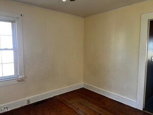 An empty room with cream-colored walls and a wooden floor. There is a window on the left side letting in natural light. The ceiling and baseboards are white, and there is an open doorway on the right. The walls and floor show signs of wear.