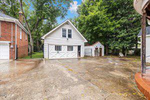 A white, two-story detached garage with a pitched roof and large doors is surrounded by trees. The building stands on a spacious concrete driveway, with a small shed in the background and a red-brick house to the left. The sky is partly cloudy.