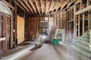 Interior of an abandoned building with exposed wooden beams and unfinished walls. A boarded-up window, scattered debris, a green plastic bin, and a staircase are visible. Sunlight enters through gaps, illuminating the dusty floor.