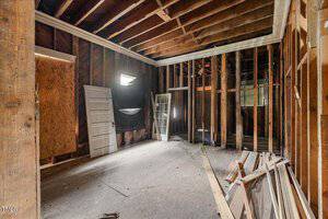 An unfinished room with exposed wooden framing on the walls and ceiling. Several doors and wooden panels are leaning against the walls. A small window provides limited light, and construction materials are scattered on the floor.